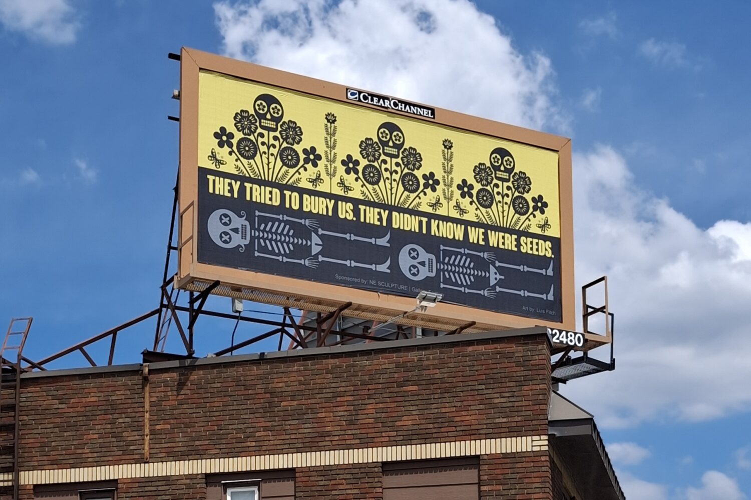 A large roadside billboard displays the words, "They Tried to Bury Us. They Didn't Know We Were Seeds." The artwork is divided horizontally: below, two skeletal figures lie in a black landscape; above, colorful flowers bloom beneath a bright yellow sky, symbolizing resilience, hope, and renewal. The billboard stands at George Floyd Square in Minneapolis as part of the Social Justice Billboard Project.