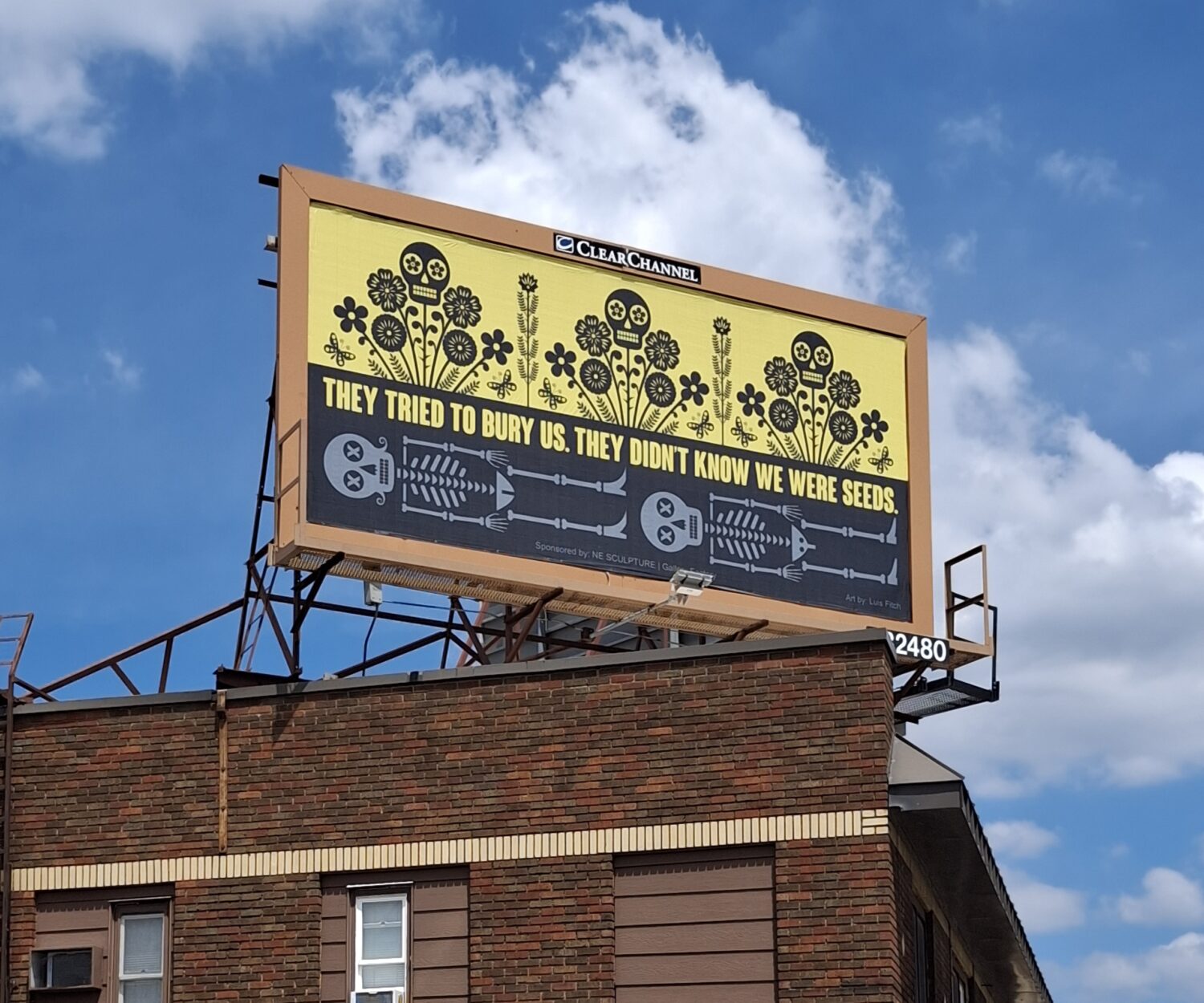 A large roadside billboard displays the words, "They Tried to Bury Us. They Didn't Know We Were Seeds." The artwork is divided horizontally: below, two skeletal figures lie in a black landscape; above, colorful flowers bloom beneath a bright yellow sky, symbolizing resilience, hope, and renewal. The billboard stands at George Floyd Square in Minneapolis as part of the Social Justice Billboard Project.
